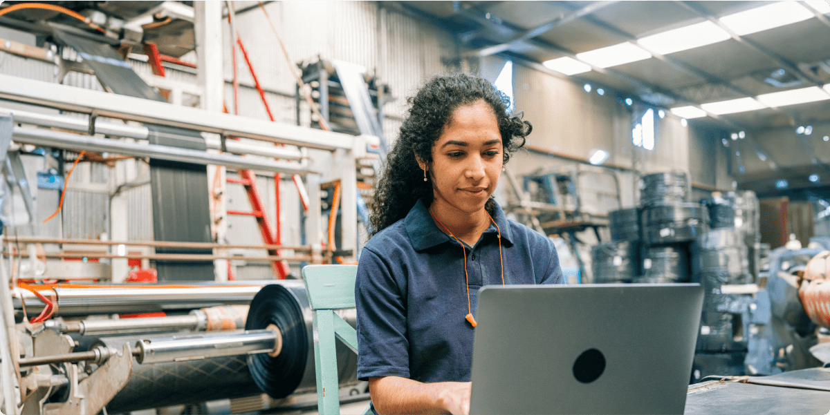 Een vrouw in een werkplaats met apparatuur en gereedschap, op haar laptop.